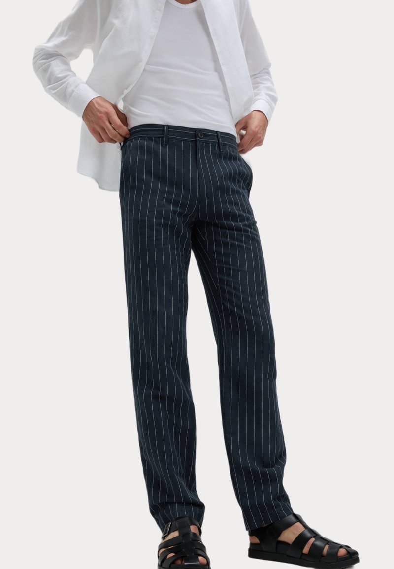 Pantalon bleu foncé à rayures, en tissu, avec une coupe droite, des poches avant et une fermeture à bouton, associé à des sandales noires.