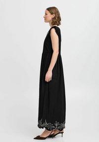Robe maxi noire avec des manches courtes à volants, taille froncée et broderie décorative de feuilles blanches le long de l'ourlet. Fabriquée en tissu léger.