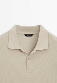 Beige polo shirt made of textured fabric, featuring a ribbed collar, three-button placket, and a subtle brand label inside the neckline.
