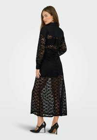Abito in pizzo nero con maniche lunghe, gonna semi-trasparente e corpetto nero solido. Abbinato a scarpe con tacchi alti nere.