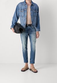 Persona vistiendo una chaqueta de mezclilla azul, una camisa de canalé beige, jeans azules deslavados, sandalias beige, sosteniendo una chaqueta de cuero negro plegada contra un fondo blanco.