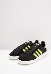 Svarta mockaskor med tre djärva neongula ränder, vit gummisula, svarta snören och en märkesdetalj på tungan.