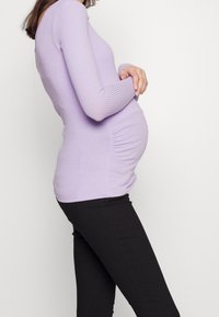 Top de maternidad de canalé en color lila con mangas largas, que presenta detalles de fruncido a los lados, combinado con pantalones negros ajustados.