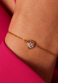Gouden armband met een hartvormige sluiting, voorzien van een heldere edelsteen in het midden. Kettingontwerp met een gladde textuur tegen de huid. Roze stoffen achtergrond.