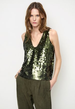 Jonge vrouw met lichtbruin haar, die een mouwloze groene paillettentop en donkergroene broek draagt, met handen in de zakken.