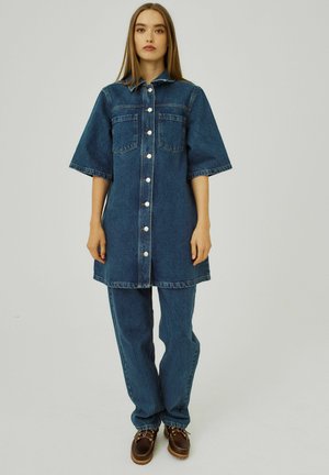 Denim-mekko lyhyillä hihoilla, edessä nappilukitus, kaksi rintataskua ja rento malli, yhdistettynä yhteensopiviin sinisiin denim-housuihin ja ruskeisiin kenkiin.