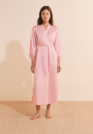 Femme debout pieds nus portant une longue robe en satin rose avec une bordure en dentelle aux manches nouée à la taille sur un fond neutre.