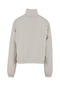 Lys grå, glidelåsjakke med høy krage, elastiske mansjetter og fald. Har hvite kanter langs skuldrene. Glatt tekstur, minimalistisk design.