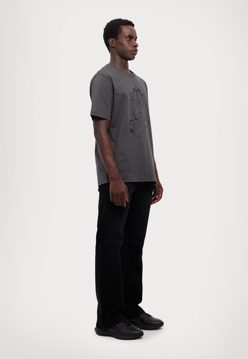 T-shirt gris en coton avec un imprime graphique noir d'un chat cartoon, associé à un pantalon noir et des chaussures noires, présenté en position debout.