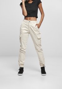 Persona vestida con pantalones cargo beige, zapatillas altas negras y un top negro de botones corto, de pie contra un fondo claro y liso.