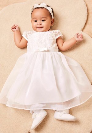 Weißes Kleid mit floralem Stickerei, transparenter Überlagerung und einer Satin-Schleife in der Taille. Weiche weiße Schuhe mit Spitze-Details. Heller Hintergrund.