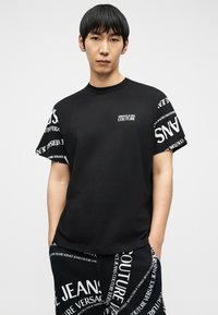 Νεαρός άνδρας που φοράει μαύρο T-shirt με στρογγυλή λαιμόκοψη και ταιριαστό παντελόνι με λευκά μοτίβα κειμένου "Versace Jeans Couture", με τα χέρια στις τσέπες.