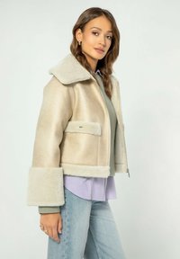 Giacca in shearling beige con ampio colletto, chiusura a zip, due tasche frontali e rifiniture in pelliccia a contrasto su polsini e orlo.