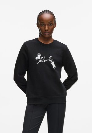 Zwarte sweatshirt van zacht materiaal met een grafische afbeelding van twee katten en het woord "Karl" in witte tekst. Klassiek model met ronde hals.