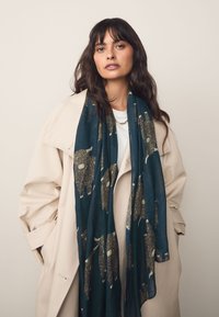 Sciarpa turchese scuro con motivo ripetuto di bisonti illustrati e testurizzati. Indossata con un trench color beige chiaro. Tessuto morbido con design fluente.