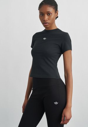 Kvinde står mod grå baggrund iført tætsiddende sort Adidas t-shirt og leggings med hvide Adidas-logoer på bryst og lår.