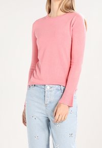 Vaaleanpunainen pitkähihaiset neule, jossa on pyöreä pääntie ja ribbauskä wrists, yhdistettynä vaaleansinisiin farkkuihin, joissa on brodeerattuja kukkakoristeita.