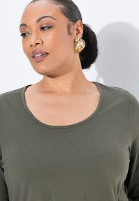 Oliivinvihreä pitkähihainen paita pyöreällä pääntiellä, jossa on sileä pinta; mallilla on päällään suurikokoinen kultaketju korusteena.