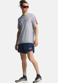 Osoba koja nosi sivi Adidas Berlin Marathon majicu, tamnoplave šortseve, crno-bijele tenisice i sportske naočale s crvenim staklima stoji bočno.