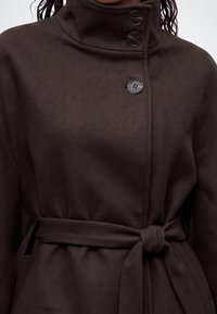 Manteau marron foncé à col montant, fermé par trois boutons et une ceinture assortie nouée à la taille, porté par une personne aux cheveux tressés.