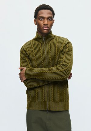 Junger Mann mit kurzen lockigen Haaren, der einen grünen Strickpullover mit Reißverschluss und passende grüne Hosen trägt, steht mit verschränkten Armen.