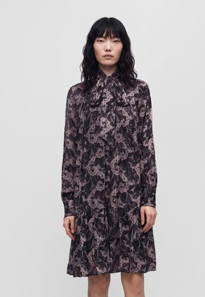 Une femme porte une robe sombre à imprimé floral, ornée d’un nœud au cou, sur un fond clair et uni.