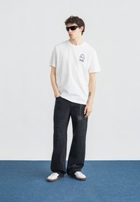 Νέος άνδρας με μαύρα γυαλιά ηλίου, λευκό T-shirt Puma, μαύρο τζιν με ξεβαμμένο εφέ και διακοσμητικά με καρφιά, και λευκά αθλητικά παπούτσια, στέκεται πάνω σε μπλε χαλί.