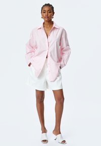 Light pink oversized shirt with white embroidered patterns, button-up front, and long sleeves. Paired with white shorts and open-toe mules.