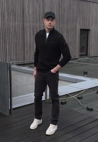 Homme portant une casquette noire, un pull noir et un pantalon avec des baskets blanches, debout sur une terrasse sur le toit près d'un mur en bois et d'une rampe en verre.
