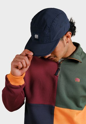 Granatowa czapka z logo z przodu, noszona nisko, w połączeniu z zapinaną na zamek, wielokolorową bluzą z sekcjami w odcieniach maroon, zielonym, granatowym i pomarańczowym.