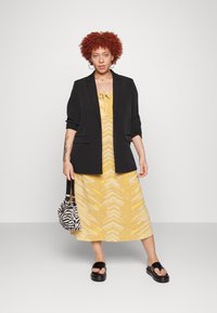 Blazer preto sobre um vestido amarelo com um padrão de zigzag, sandálias pretas e uma bolsa listrada. Cabelo castanho encaracolado e joias minimalistas.