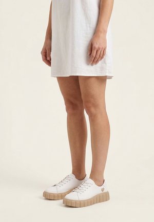 Person iført hvide lave sneakers med beige såler og et lille hjerte-detalje, kombineret med en hvid kort kjole.