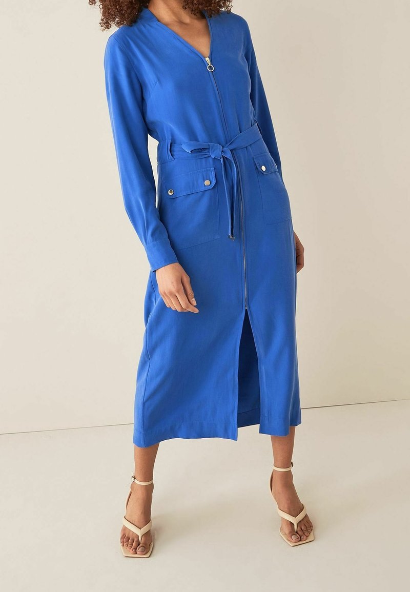 Langwerpige blauwe jurk met een ritssluiting aan de voorkant, een ceintuur om de taille en twee borstzakken. Glad materiaal met een lengte tot halverwege de kuit en een splitdetail.