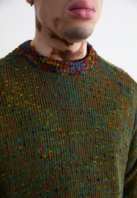 Textured green sweater with multicolored specks, featuring a ribbed neckline and loose-knit design. Patterns include small, vibrant accents.