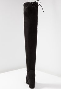 Bottes noires montant au-dessus du genou en daim doux, avec un talon bloc et un détail noué en haut. Texture lisse et design ajusté.