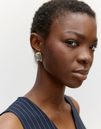 Silberne, metallische Ohrringe mit einer organischen Form, die sanfte Kurven aufweisen. Das Modell trägt ein ärmelloses Oberteil mit navyblauen Streifen. Neutraler Hintergrund.