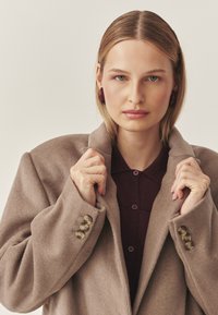 Beiger Wollmantel mit großen Revers, mit drei Akzentknöpfen am Ärmel. Getragen über einem dunkelburgunderfarbenen Hemd mit Kragen. Glatte Textur.