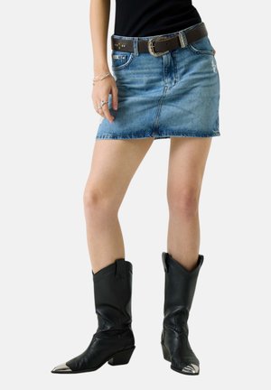 Donna che indossa una minigonna in denim blu con tasche frontali, una cintura nera, un top nero, anelli e braccialetti d'argento e stivali da cowboy neri con punte in metallo.