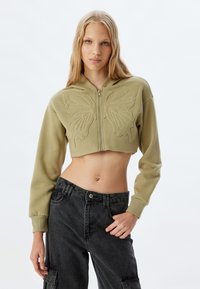 Sudadera recortada de color verde oliva con bordado de mariposa, cierre de cremallera en la parte delantera, mangas largas y un acabado texturizado. Combinada con pantalones cargo oscuros.