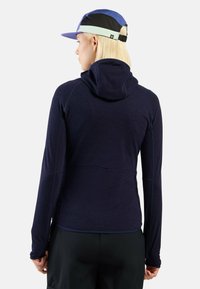 Marineblauer Hoodie mit langen Ärmeln und taillierter Passform, strukturiertem Stoff und enganliegender Kapuze. Das Model trägt eine mehrfarbige Mütze.