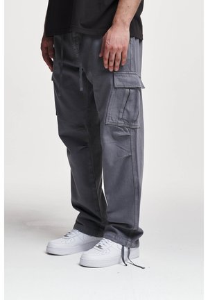 Mann trägt graue Cargohose mit Seitentaschen und weiße Sneaker, steht vor einem einfarbigen hellen Hintergrund.