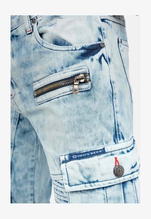 Pantaloni cargo in denim azzurro chiaro con un aspetto sbiadito e strappato. Presentano una tasca con zip e una patch con logo accentuata da cuciture rosse.