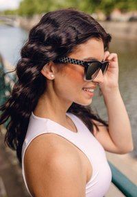 Sluneční brýle s černým vzorovaným rámem a tmavými skly, s průhledným dolním okrajem. Modelka má na sobě bílé tílko bez rukávů.