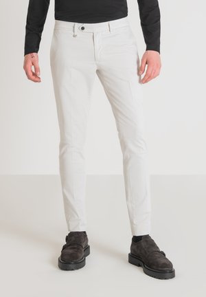 Pantaloni bianchi slim fit in cotone, con chiusura a bottone, tasche laterali e una superficie liscia. Abbinati a scarpe marroni scure.