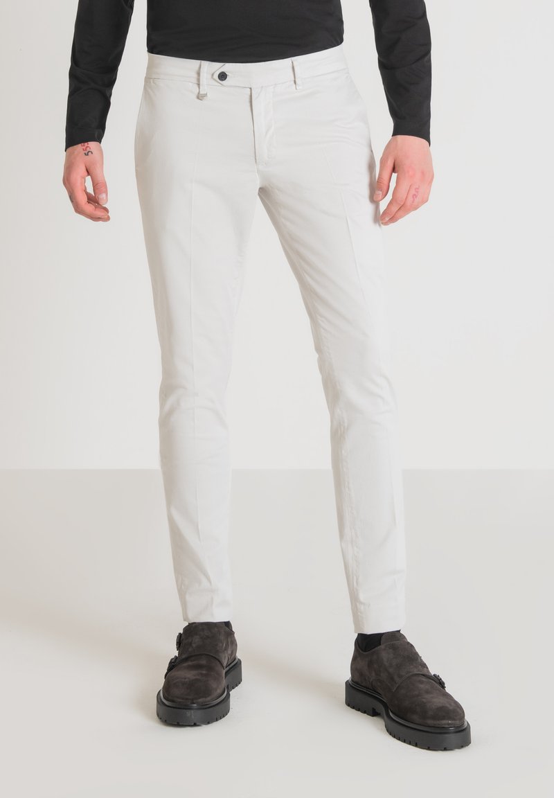 Pantaloni bianchi slim fit in cotone, con chiusura a bottone, tasche laterali e una superficie liscia. Abbinati a scarpe marroni scure.