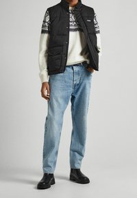 Musta toppaliivi valkoisen neulepuseron päällä, jossa tummia geometrisia kuvioita, yhdistetty vaaleansinisiin farkkuihin ja mustiin nilkkureihin.