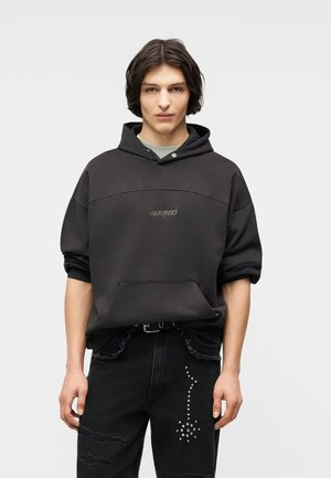 Νεαρό άτομο με μεσαίου μήκους σκούρα μαλλιά που φοράει μαύρη κουκούλα με την επιγραφή "wasted" και μαύρο τζιν με διακοσμητικό σχέδιο με ανθάκια που μοιάζουν με πουά.