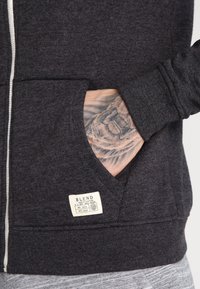 Dunkelgrauer Hoodie mit Reißverschluss, texturiertem Stoff, Seitentaschen und einem Etikett mit der Aufschrift "BLEND." Besitzt ein lässiges Design und gerippte Bündchen.