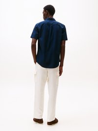 Homem vestido com uma camisa de mangas curtas azul-marinho, calças cargo brancas e mocassins castanhos, de pé, virado de costas, num fundo branco liso.