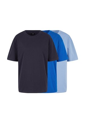 Jeune homme aux cheveux courts portant un t-shirt oversize bleu vif uni et un pantalon sombre, debout devant un fond gris clair.